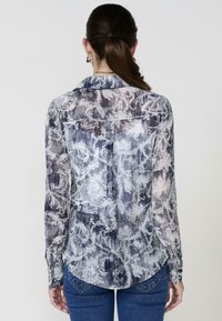 Una donna con capelli lunghi indossa una camicia a maniche lunghe con un motivo e dei jeans, girata di spalle contro uno sfondo neutro.
