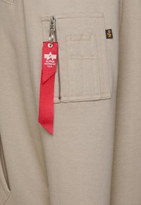Bežna dukserica z zadrgo in žepom, ki ima rdečo tkano oznako z belim logotipom ter teksturirano blago z ojačanimi šivi.