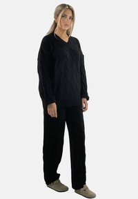 Schwarzer Strickpullover mit V-Ausschnitt und Zopfmuster, kombiniert mit passenden schwarzen Hosen. Die Schuhe sind hell und weich.