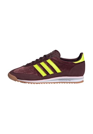 Niski profil butów sportowych Adidas w ciemnym bordowym kolorze z jasnymi żółtymi paskami i pętelką na pięcie, gumową podeszwą i białą śródpodeszwą z logo Adidas.
