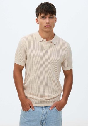 Joven con cabello corto y oscuro que lleva una camiseta polo beige de punto de manga corta y vaqueros azul claro con las manos en los bolsillos.