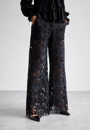 Kvinde iført brede bukser med sort blomstret blonder og matchende top med hænderne i lommerne, stående på gråt gulv.