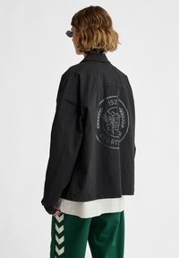 Svart oversized jacka med sänkta axlar, prydd med en stor grafisk design på baksidan med en bi och lagerblad. Bärs över en vit skjorta.