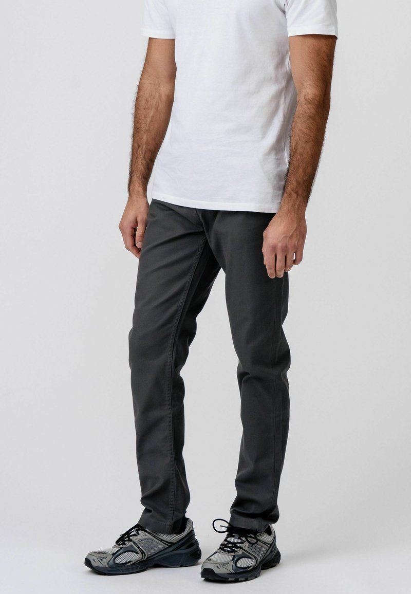 Homme portant un pantalon gris foncé, une chemise blanche à manches courtes et des chaussures de sport grises et noires, debout devant un fond uni.