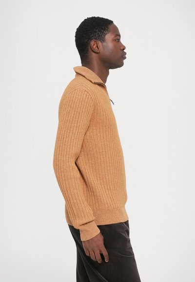 Ribana, svetlo rjava pletena jopica s ovratnikom in četrtinskim zadrgotom. V kombinaciji s temnimi kordnimi hlačami, ki prikazujejo oprijet silhueto.