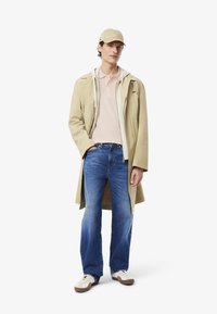 Beige lång kappa över en vit huvtröja och en rosa polo, blå jeans och en beige baseballkeps. Bruna och vita sneakers fulländar outfiten.