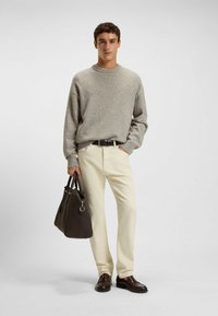Maglione grigio a maglia, pantaloni crema, cintura in pelle marrone, scarpe abbinate e una borsa in pelle marrone scuro. Il modello sta in piedi su uno sfondo neutro.