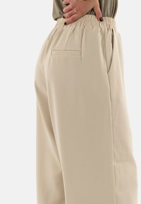 Pantalons beige à taille élastique avec poches latérales ; texture lisse, design à jambes larges et légères plis à la taille pour plus de confort.