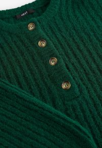 Groene geribde trui met vijf bruine knopen aan de voorkant. Gemaakt van een textuurstof, die een zachte, fuzzy uitstraling heeft.