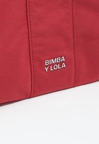 Bolsa de tela roja con una textura suave. Presenta un logo plateado que dice "BIMBA Y LOLA." Costuras visibles a lo largo de los bordes.
