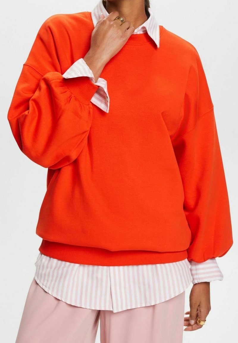 Sudadera naranja de gran tamaño con mangas globo, que presenta una camisa de cuello blanca y rosa a rayas debajo. Textura de tela suave, diseño casual.
