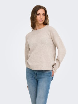 Jeune femme aux cheveux ondulés portant un pull beige et un jean bleu, debout avec une main dans la poche devant un fond clair.