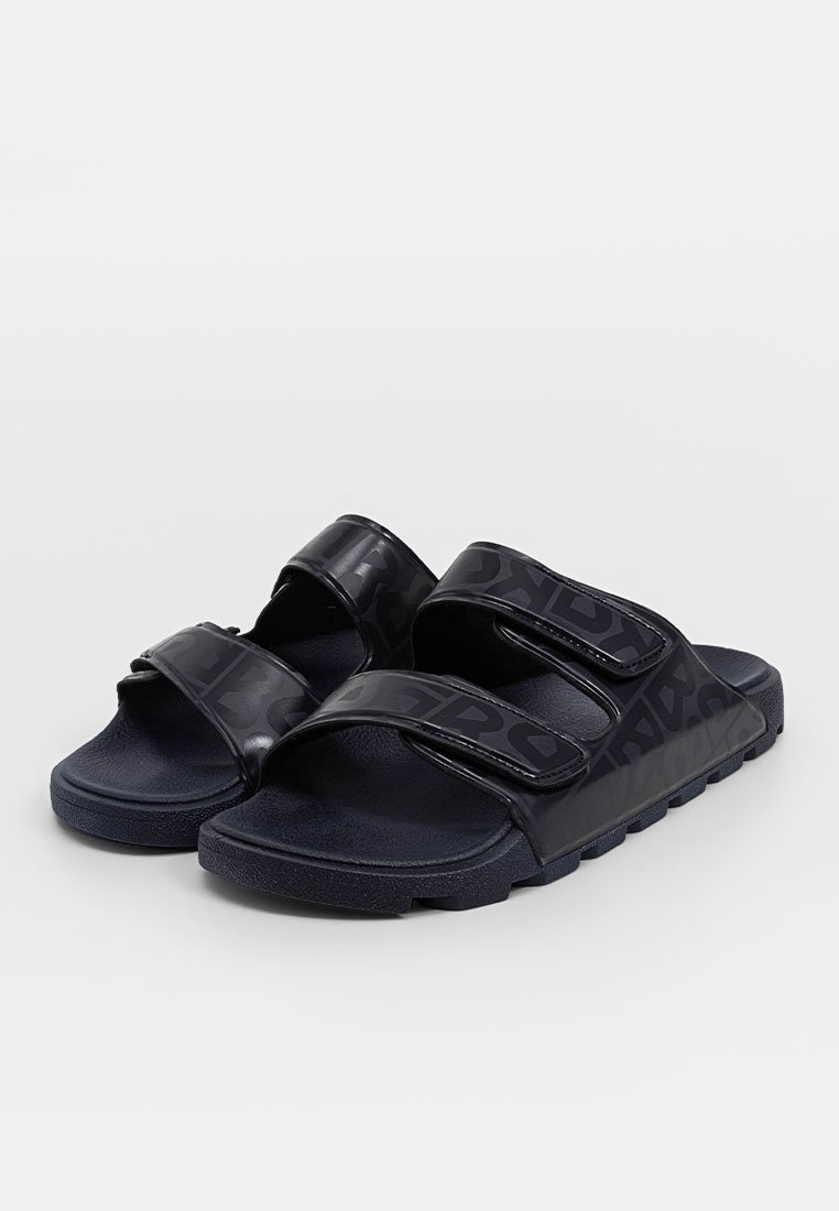 Schwarze Riemchensandalen mit zwei verstellbaren Riemen, die eine glänzende Textur und ein geprägtes Logo-Muster aufweisen. Gummisohle für einen guten Halt.
