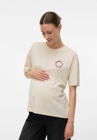 Zwangere vrouw in een beige shirt met rode tekst, zachtjes haar buik vasthoudend, staande tegen een effen witte achtergrond.