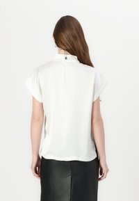 Blouse blanche à manches courtes avec un col haut, comportant une fermeture arrière en forme de trou de serrure et en tissu lisse et léger. Associée à une jupe noire.