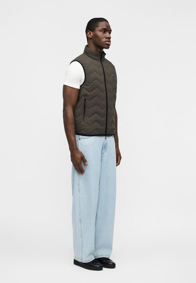 Άνδρας που φοράει ανοιχτό μπλε τζιν παντελόνι με φαρδιά μπατζάκια, λευκό κοντομάνικο πουκάμισο και σκουροπράσινο αμάνικο μπουφάν με φερμουάρ και μαύρα παπούτσια.