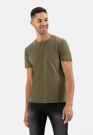 Olivgrünes, kurzärmliges Henley-Shirt mit Rundhalsausschnitt, zwei schwarzen Knöpfen und einer feinen Textur. Kombiniert mit dunklen Jeans.