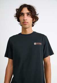 Sort bomulds T-shirt med korte ærmer, der har et lille rød og hvidt "Converse" logo-lap på venstre brystområde.