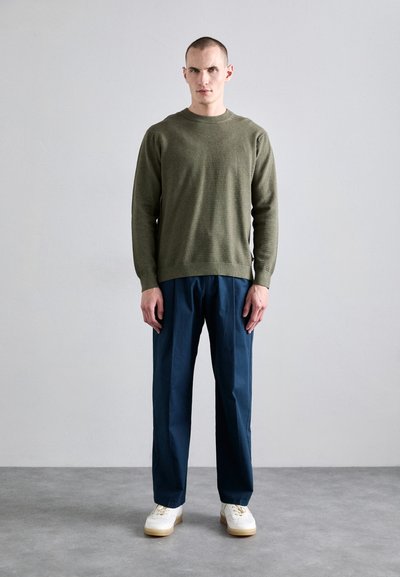 Sweater texturé vert olive, pantalon bleu marine à coupe ample, baskets blanches avec semelles claires. Design minimaliste, palette de couleurs neutres.
