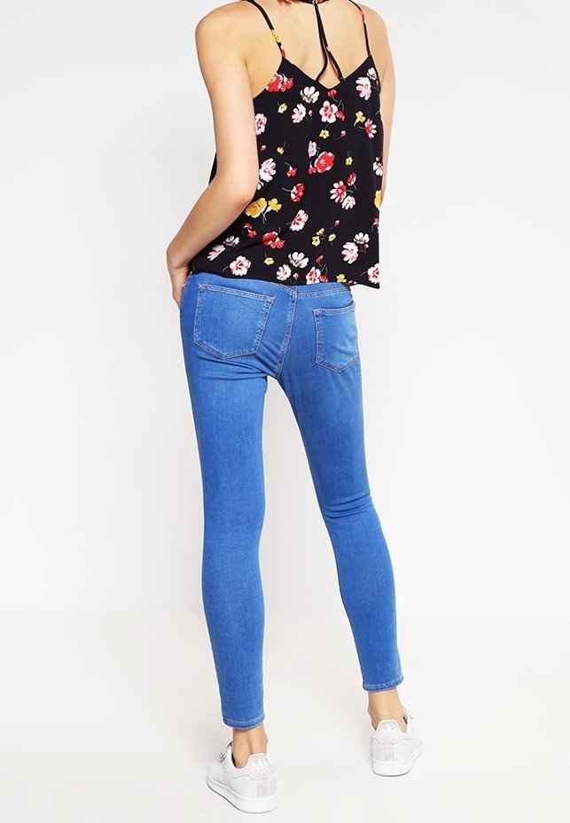 Blauwe skinny jeans met een gladde textuur en een nauwsluitend ontwerp, gecombineerd met een zwarte bloemenblouse met rode en gele bloemen.