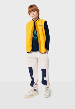 Chaleco de forro polar amarillo con botones de presión plateados, camisa azul marino con estampado gráfico, pantalones beige con parches azul marino y zapatillas blancas.