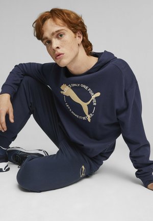 Granatowy zestaw bluz i spodni wykonany z miękkiego materiału. Bluza ma złote logo Puma oraz napis, luźny krój i kaptur.