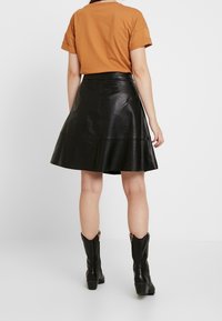 Musta nahasta valmistettu liehuva hame, jossa on sileä pinta, yhdistetty ruskeaan t-paitaan. Malli käyttää mustia nilkkureita, joissa on vetoketjut, ja seisoo valkoista taustaa vasten.