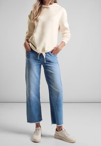 Jasno beżowa bluza z kapturem, z regulowanym sznurkiem, w połączeniu z niebieskimi spodniami dzwonami i kremowymi trampkami, o luźnym kroju i miękkiej teksturze.