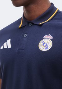 Homme portant un polo bleu marine avec un col rayé jaune, logo du Real Madrid sur la poitrine gauche et logo blanc Adidas sur la poitrine droite.
