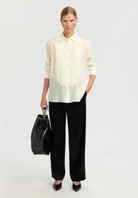 Lichtcrème overhemd met verticale plooien, gecombineerd met zwarte wijde broek. Model houdt een zwarte tote bag vast met contrasterende stiksels.