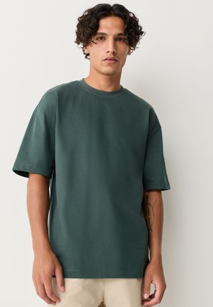 Junger Mann mit lockigem dunklem Haar, der ein dunkelgrünes übergroßes T-Shirt und beige Hose trägt, steht vor einer schlichten hellen Wand.