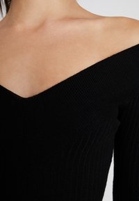 Haut noir côtelé avec un design épaules dénudées et un décolleté en V. Le tissu présente un motif texturé, renforçant la profondeur visuelle.