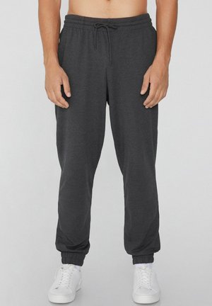 Homme portant un pantalon de jogging gris foncé avec des poignets élastiques et des baskets blanches, debout devant un fond gris clair.