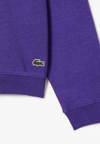 Purple sweatshirt made of soft fabric, featuring ribbed cuffs and an embroidered green crocodile logo on the left sleeve.