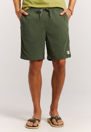 Man draagt olijfgroene short met trekkoord en zijzakken, en beige teenslippers, staand tegen een effen achtergrond.