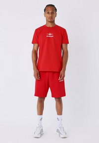 Jeune homme se tenant debout, portant un T-shirt et un short rouges assortis avec des logos blancs, des chaussettes blanches et des baskets blanches, sur un fond clair uni.