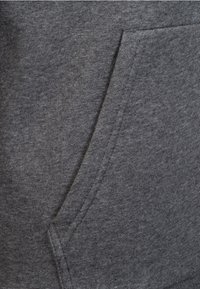Grijze hoodie stof met een kangoeroe zak. De textuur is glad met een licht opgeruwd oppervlak. De stiksels zijn zichtbaar langs de randen.
