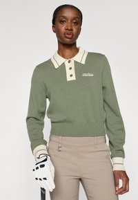 Gai zaļš adīts džemperis ar krēmkrāsas apkakli un trim pogām, ar ribotiem aprocēm un apakšmalu; uz krūtīm izšūts logo.