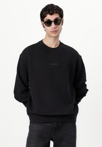 Sort crew neck sweatshirts med ribbet tekstur og et subtilt logo på brystet; parret med mørke jeans og runde solbriller.