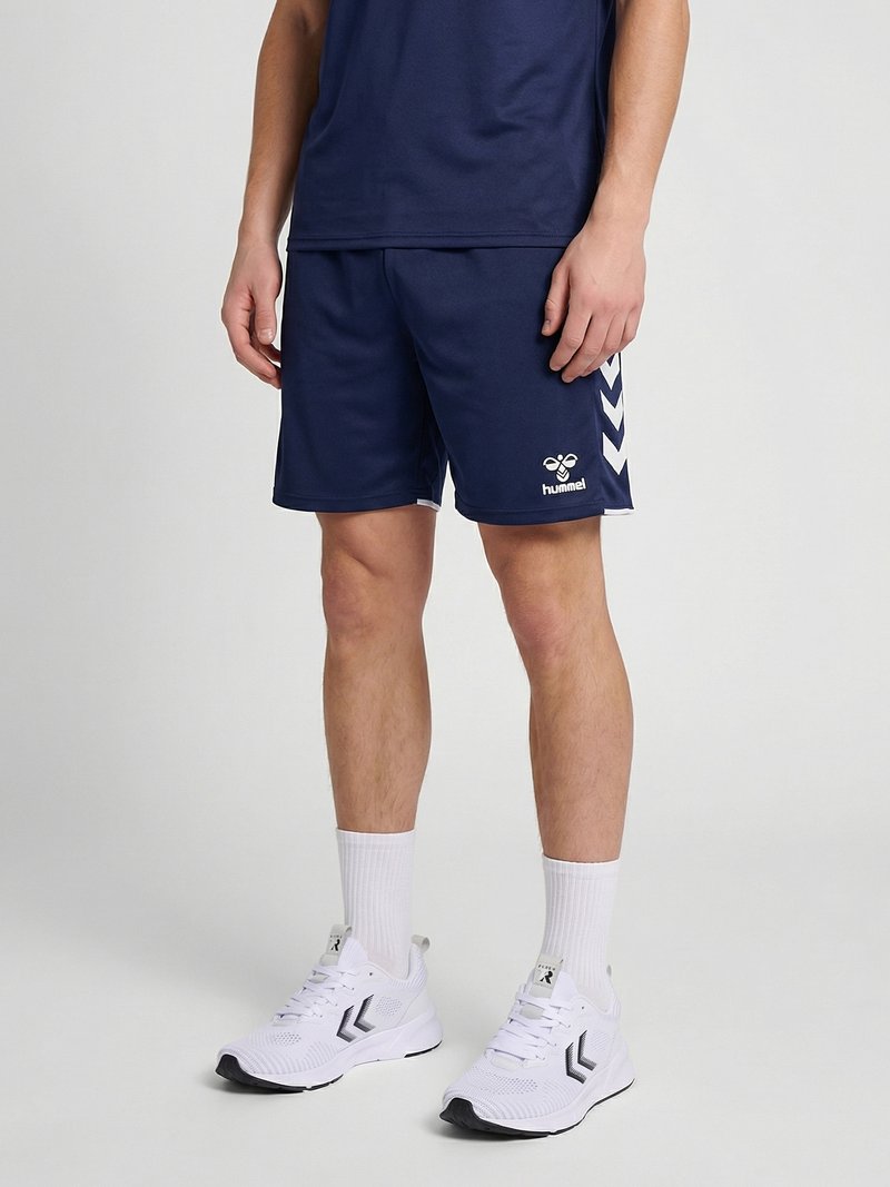 Homme portant un short de sport hummel bleu marine, des chaussettes blanches et des chaussures de sport blanches sur un fond clair uni.