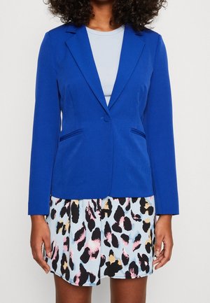 Femme portant un blazer bleu vif ajusté sur un haut bleu clair et une jupe courte avec un imprimé animalier abstrait multicolore.