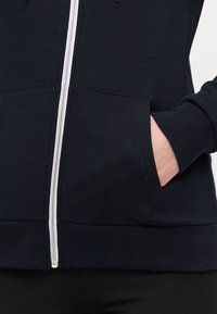 Sudadera con cremallera en color navy, confeccionada en una mezcla de algodón, que presenta una cremallera blanca, bolsillos laterales y puños y dobladillo de canalé. Textura suave, ajuste informal.