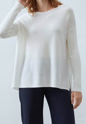 Pull blanc oversize avec des manches côtelées, un col rond et des fentes sur les côtés. Texture douce et tissu léger. Associé à un pantalon sombre.