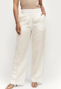 Pantaloni in lino avorio a gamba larga con tasche frontali, design a gamba dritta e dettagli di cucitura visibili, abbinati a sandali nudi con cinturini.