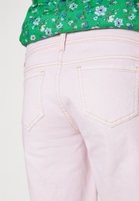 Jeans de denim rosa claro com uma textura suave, apresentando dois bolsos traseiros e costura visível numa cor contrastante. Top floral verde parcialmente visível.