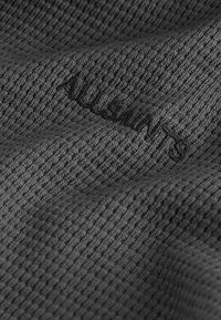 Gray textured fabric with a mesh pattern, featuring embroidered branding. The surface is detailed and slightly stretchy.