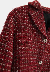 Blazer en tweed rouge texturé avec des fils noirs, blancs et dorés, présentant un col cranté et deux boutons décoratifs argentés à l'avant.