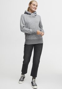 Sudadera gris con capucha y detalles de botones, con un patrón floral disperso en rosa y azul, combinada con jeans negros y zapatillas.