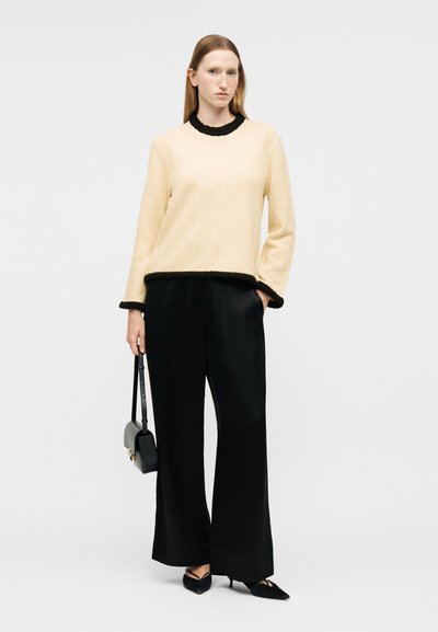 Femme aux cheveux bruns clairs et raides, portant un pull crème avec des bordures noires, un pantalon large noir, des talons noirs et tenant un sac à main noir.