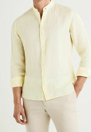 Mężczyzna ubrany w bladoróżową, długą koszulę na guziki z kołnierzem mandaryńskim, z lewą ręką w kieszeni, stojący na białym tle.